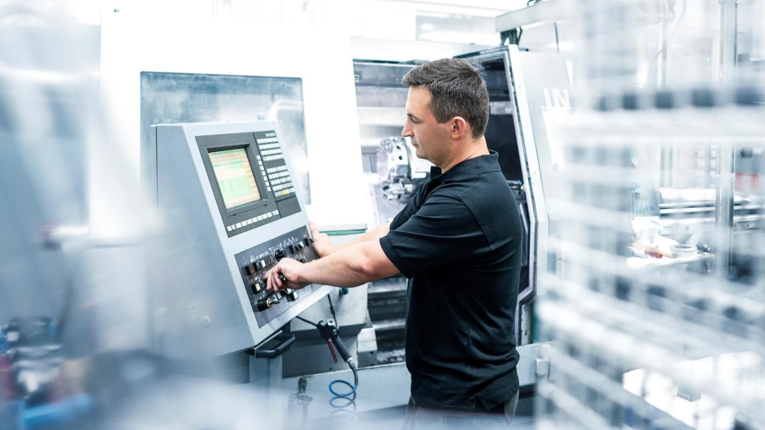 Waldemar steht neben einem Steuerungspult und bedient eine metallverarbeitenden Maschine.