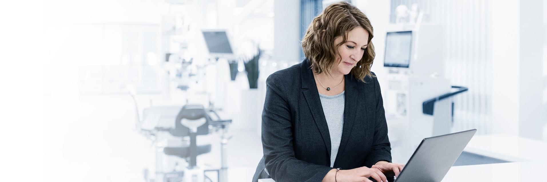Alexandra steht an einem weißen Tisch in einem modernen, hellen Raum und tippt auf einem Laptop. Sie schaut konzentriert auf den Bildschirm. Im Hintergrund sind medizinische Geräte zu sehen.