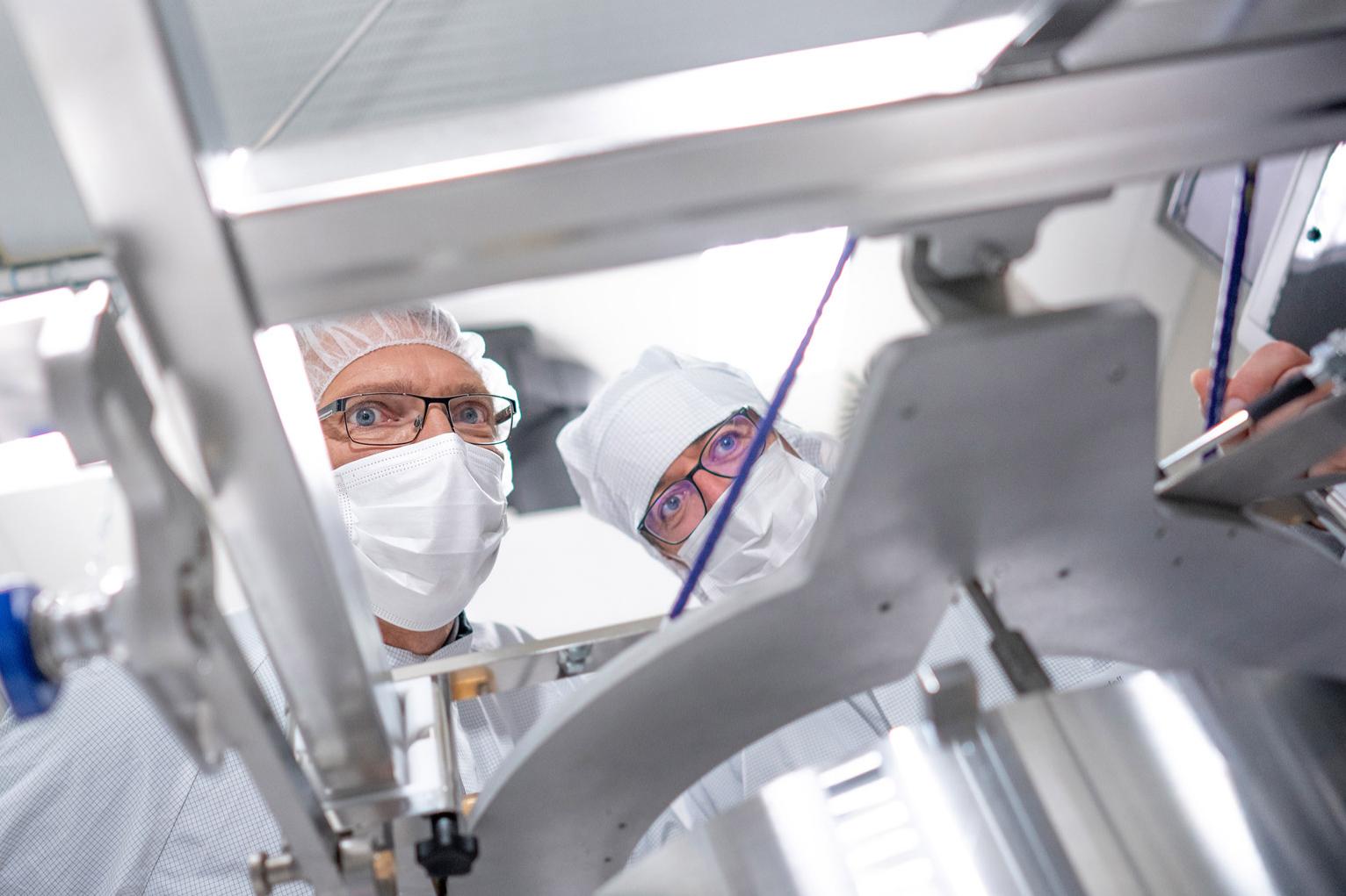 Martin and his colleague wear protective clothing and bend over a machine component for semiconductor manufacturing. They inspect it intently
