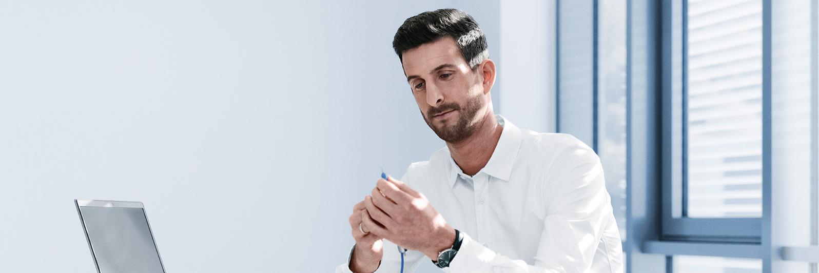 Philipp sitzt an einem weißen Schreibtisch in einem hellen Raum und hält ein Phakohandstück mit dem blauen Kabel in den Händen. Vor ihm liegt ein Laptop.