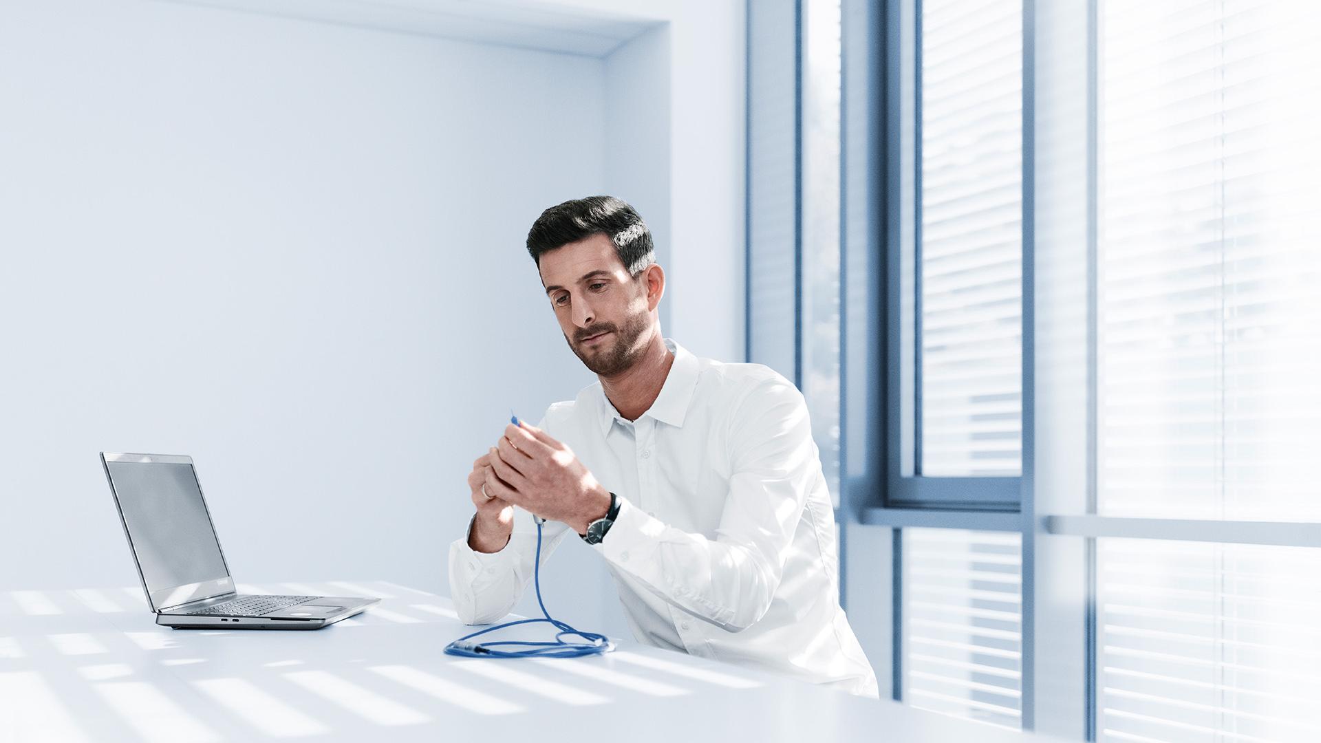 Philipp sits at a white desk in a bright room. He holds a phaco handpiece with a blue cable in his hand. There is a laptop in front of him.