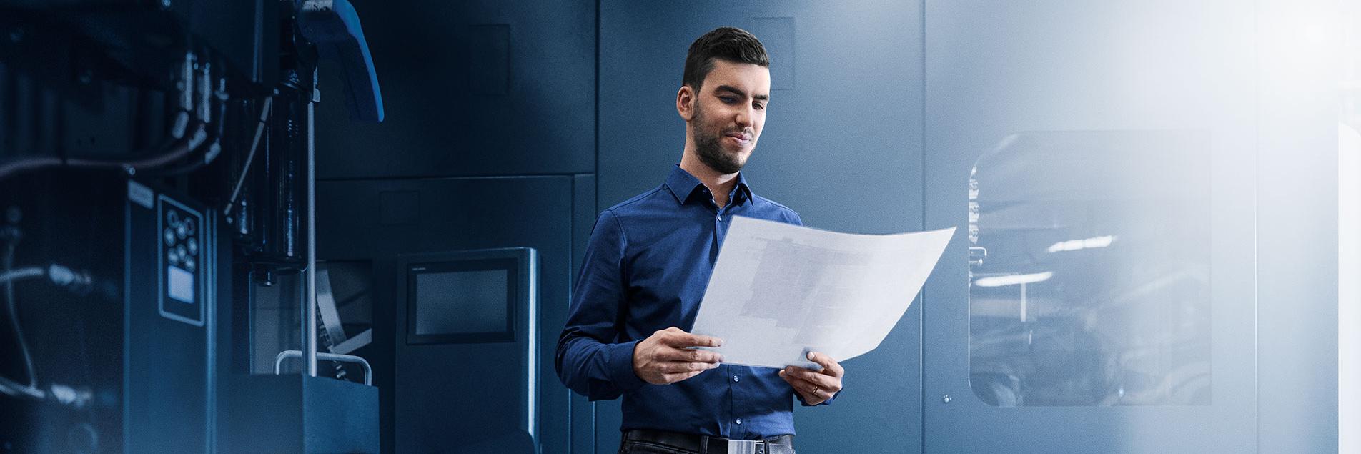A project manager stands in a dark factory environment and holds a white sheet of paper in his hand. The writing on the paper is not visible