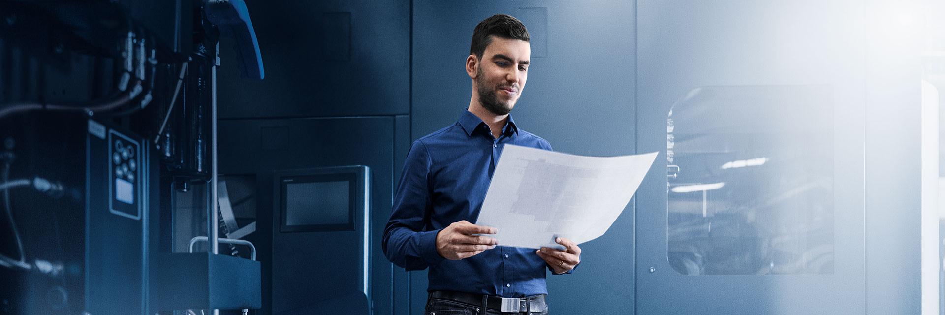 Ein Projektmanager steht in einer dunklen Fabrikumgebung und hält ein weißes Blatt Papier in der Hand, dessen Inhalt nicht sichtbar ist