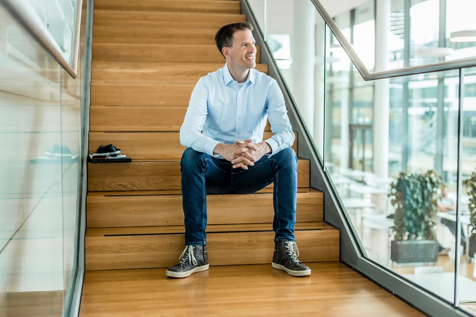 Felix sits on a wooden staircase in a modern ZEISS building, his cell phone and laptop are next to him. He is wearing a blue shirt and jeans, smiling and looking to his left.
