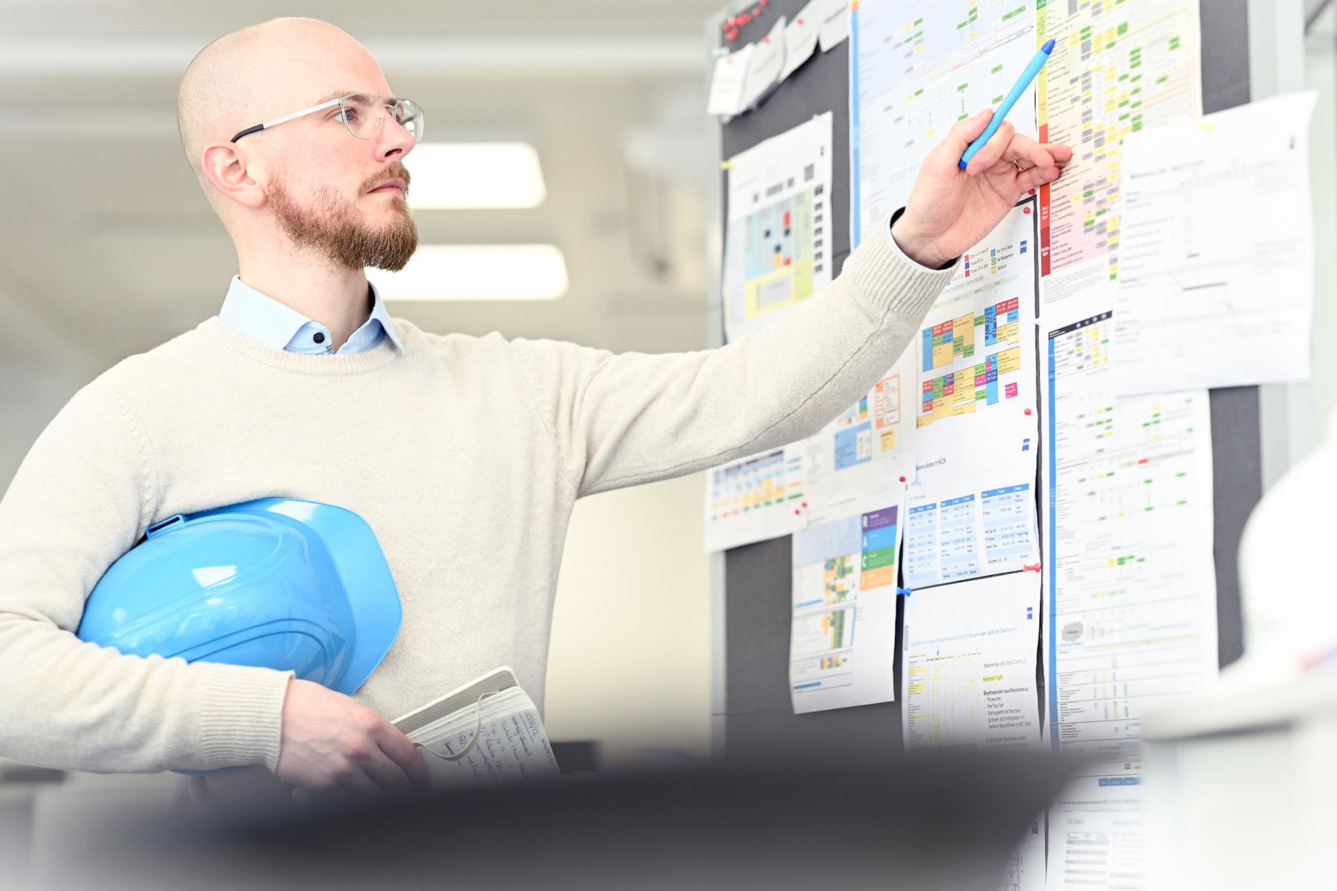 Max holds a blue hardhat under his arm and a notebook in his hand while using his pen to point at a planning board full of tables and documents.