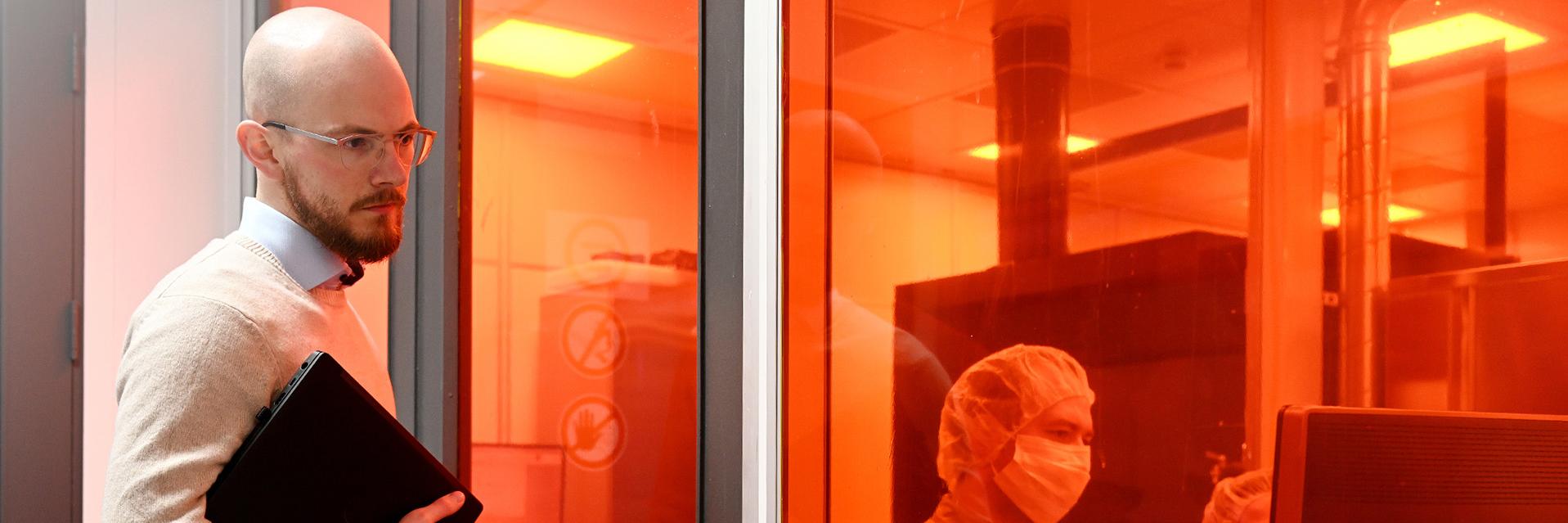 With his laptop under his arm, Max looks at the computer screen through the laboratory window, while two employees in protective clothing discuss their findings.