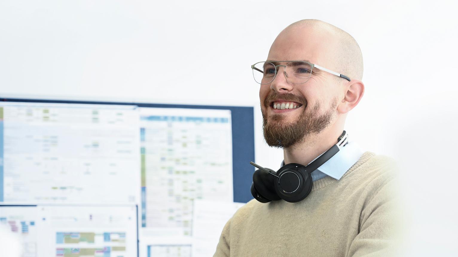 Max stands with his back to the board, headphones around his neck, as he looks ahead and smiles. He wears glasses and has a beard.