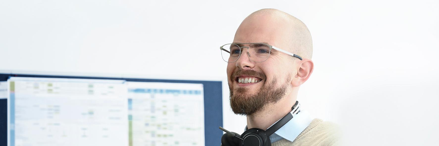 Max stands with his back to the board, headphones around his neck, as he looks ahead and smiles. He wears glasses and has a beard.