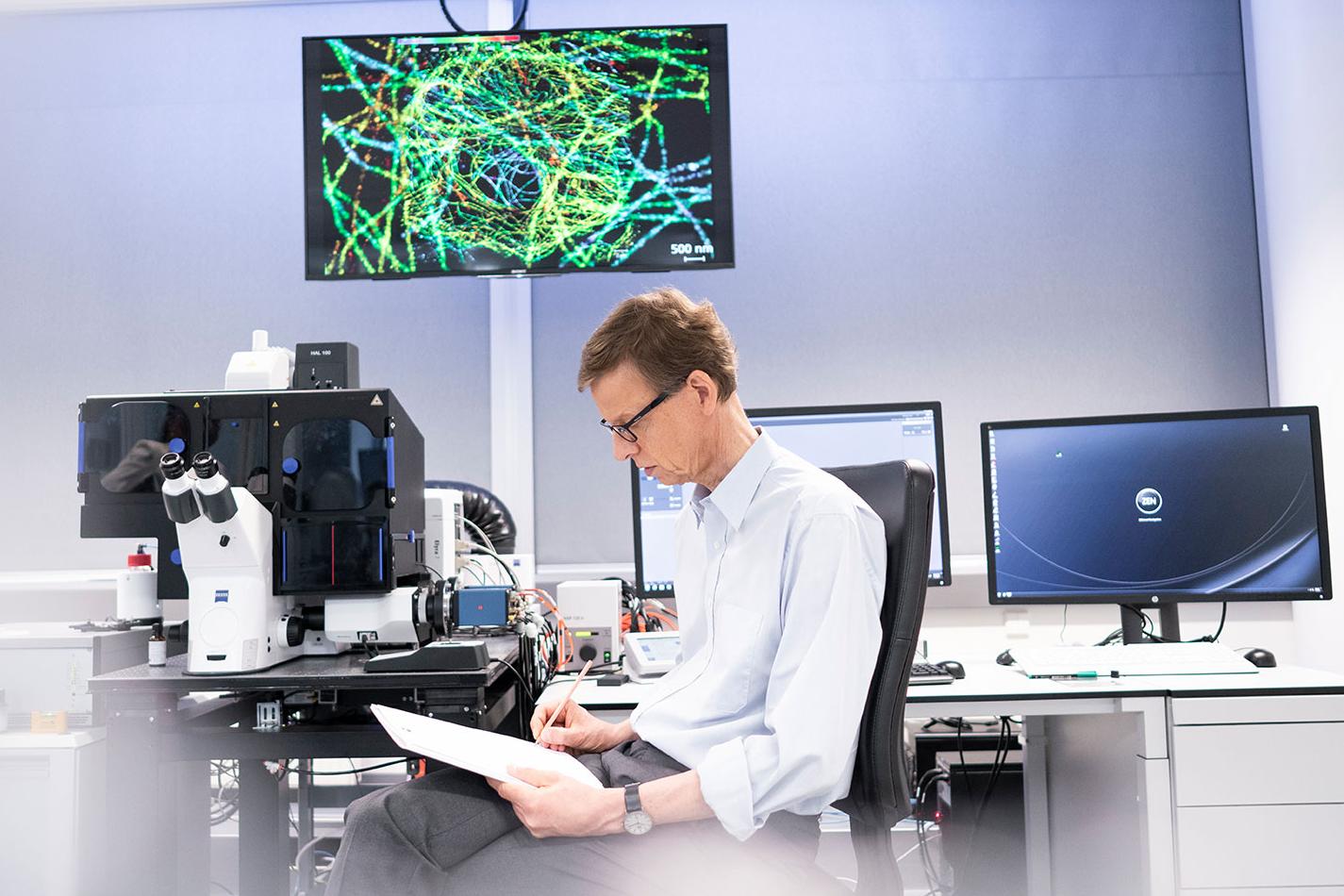 Michael sits at a desk in a ZEISS laboratory making notes on his notepad. Three monitors and a ZEISS microscope can be seen in the background.