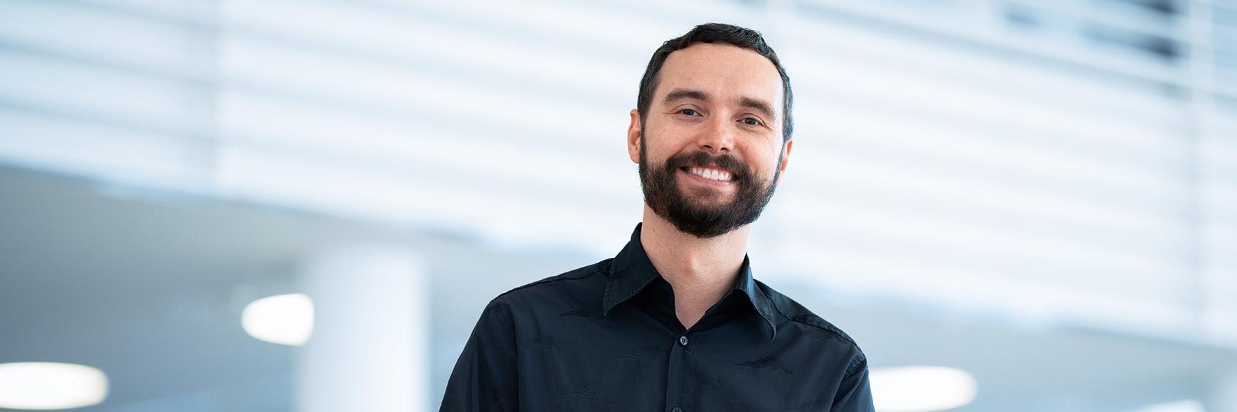 Ondrej, who wears a black shirt and has a short beard, smiles warmly at the camera, while the ZEISS office can be seen in the background.