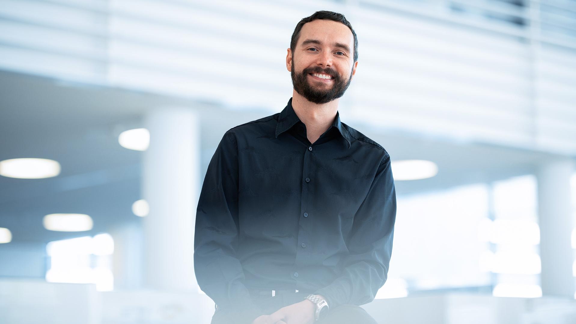Ondrej, who wears a black shirt and has a short beard, smiles warmly at the camera, while the ZEISS office can be seen in the background.