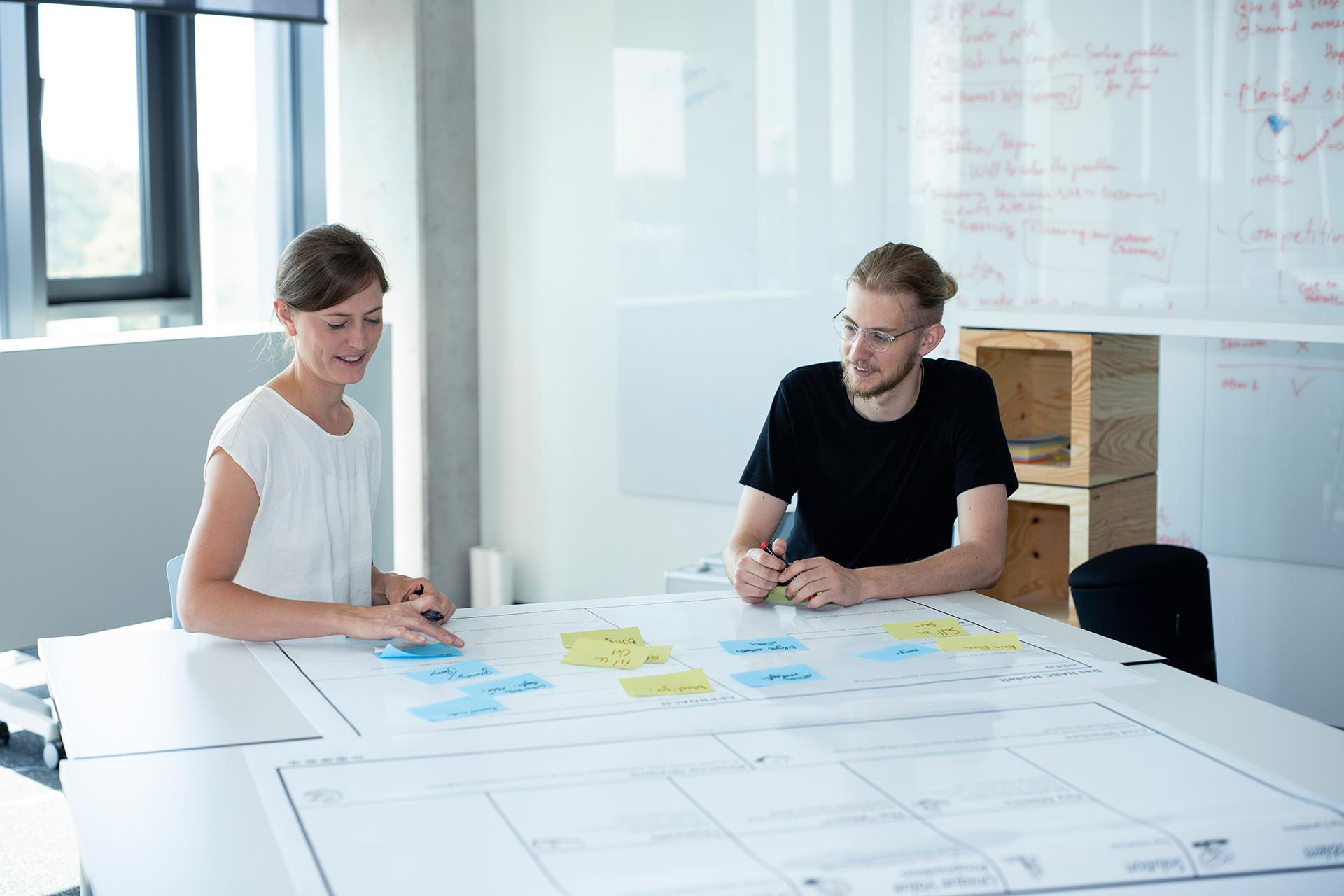 Sophia and her colleague have a discussion at the desk over sticky notes on a whiteboard.