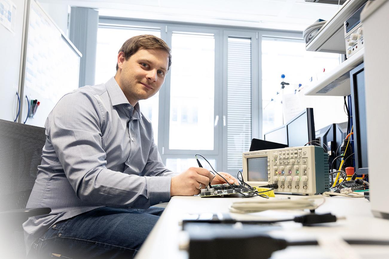 Steffen works at a desk and tests an electronic device while looking at the camera.