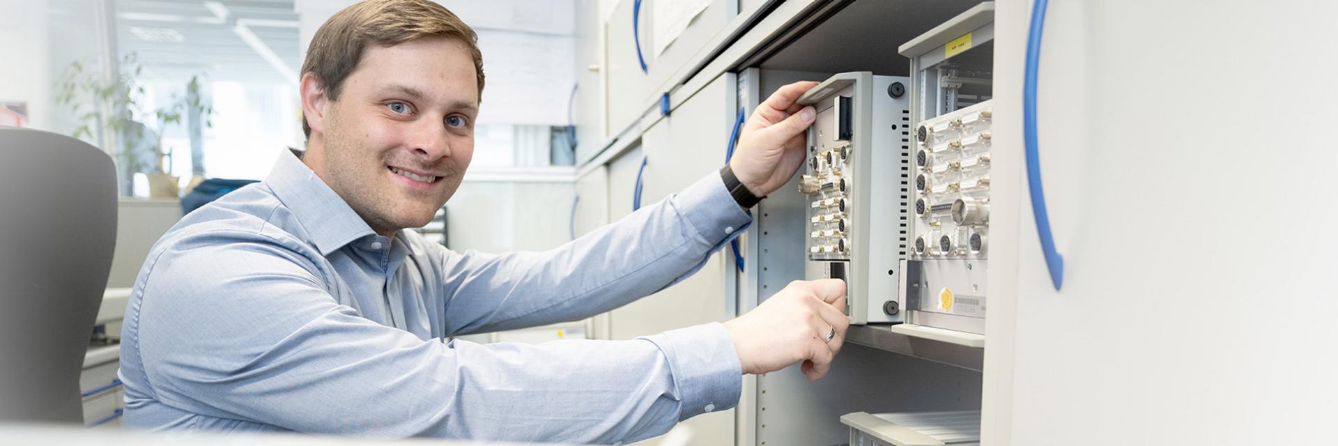 Steffen adjusts a device in a server cabinet, while looking into the camera and smiling.