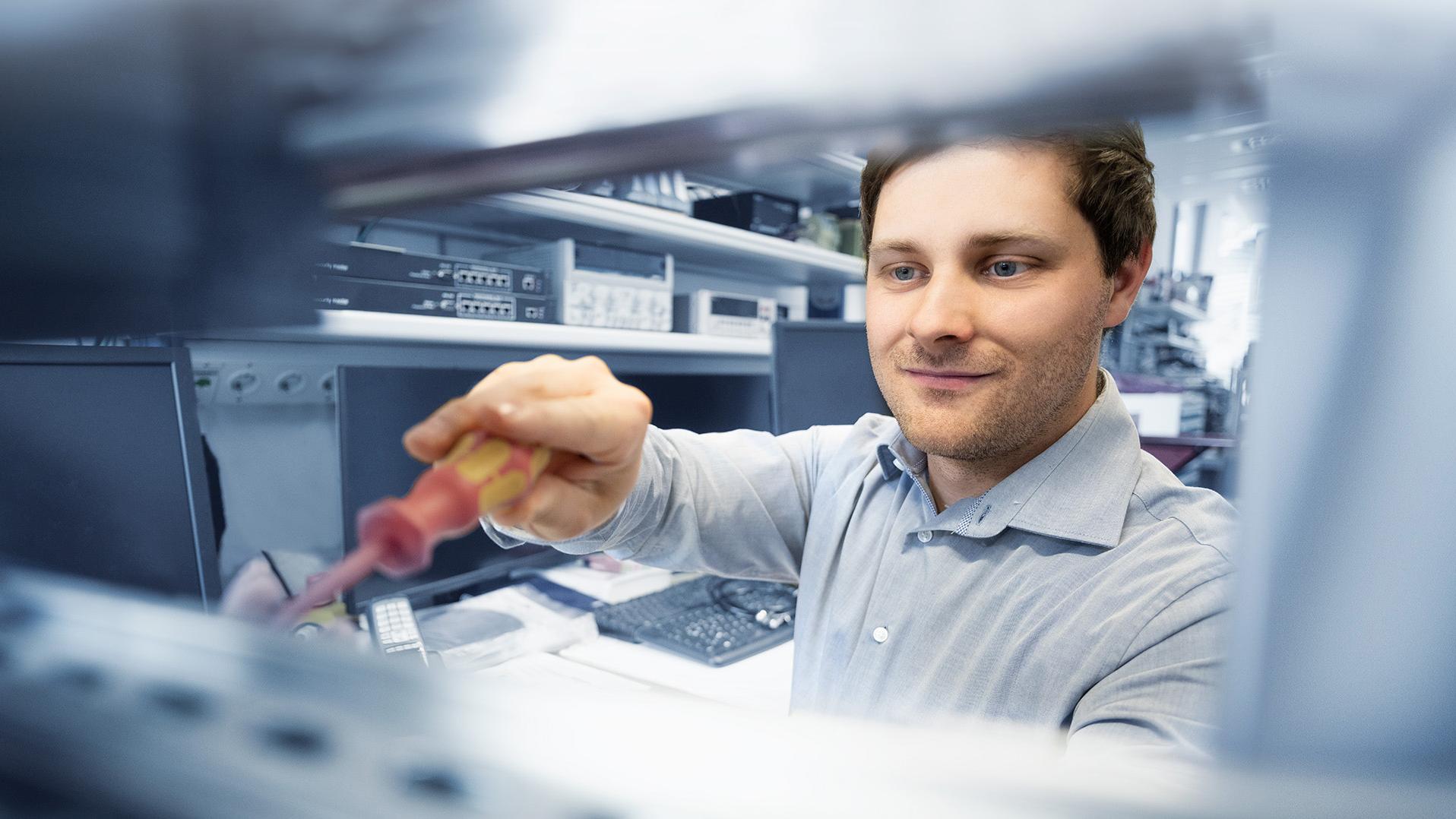 Steffen arbeitet mit einem Schraubenzieher an einem elektronischen Gerät in einem technischen Arbeitsbereich.