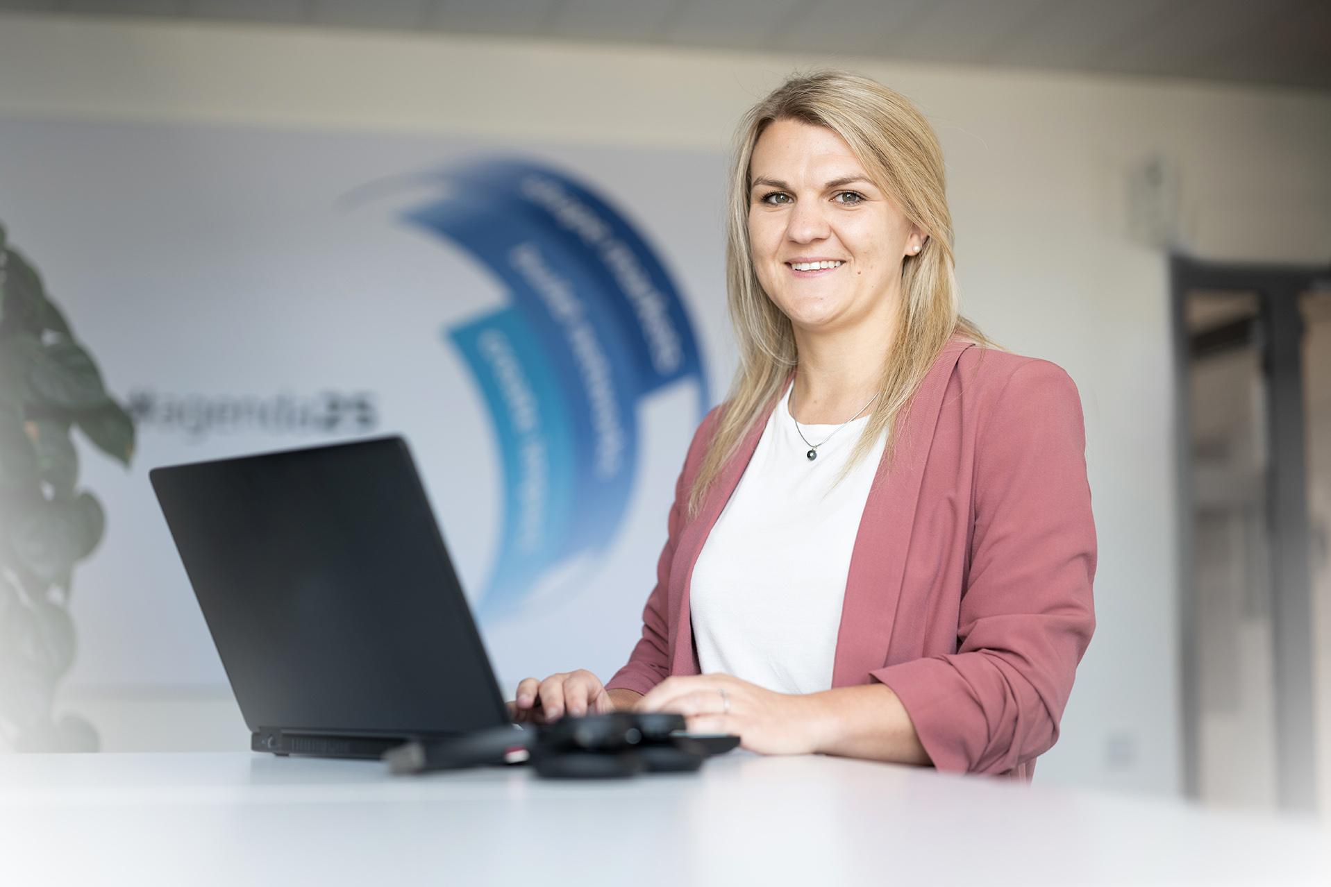 Stephanie sits at a desk, types on a laptop and smiles as she looks at the camera.