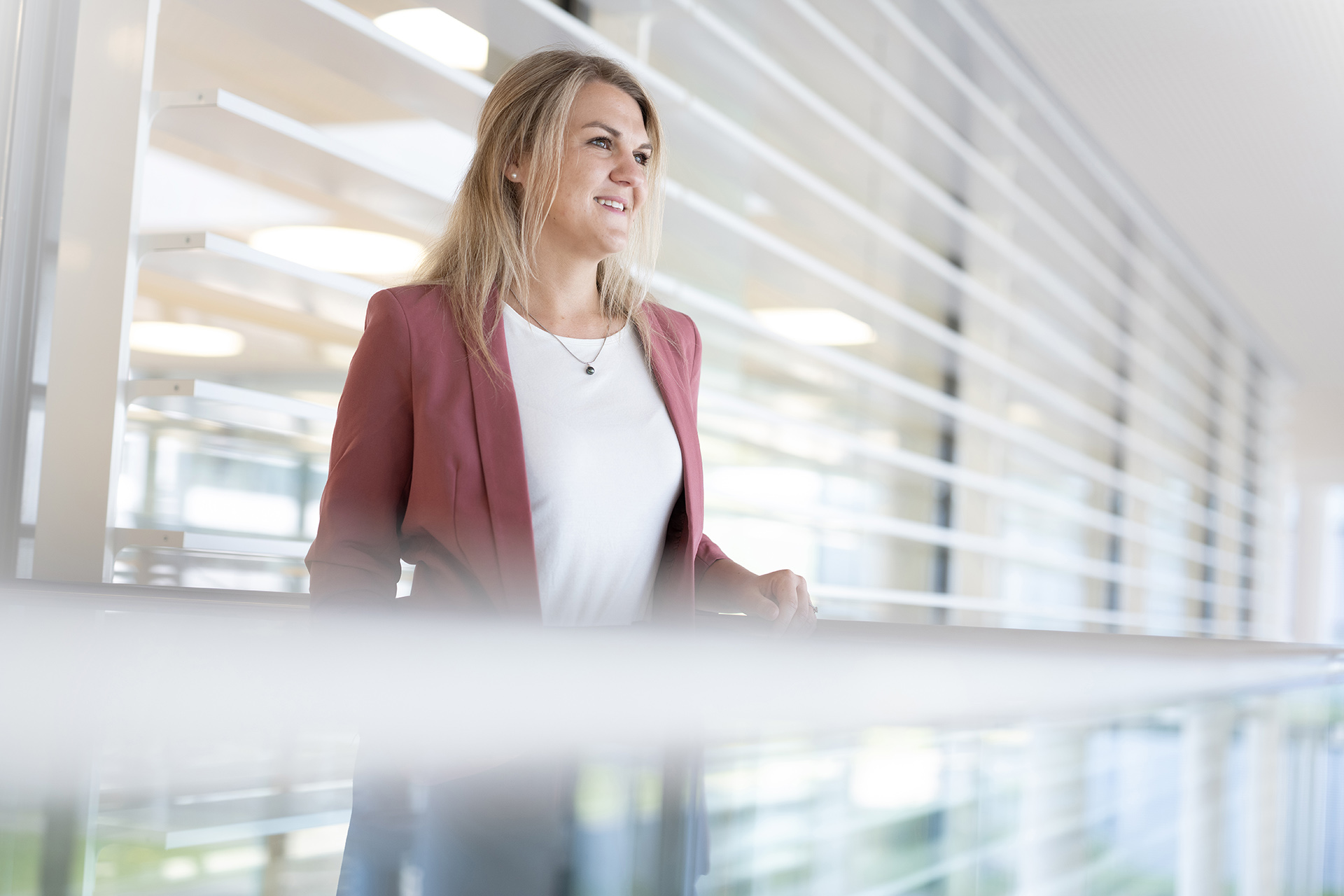 Stephanie wears a pink blazer and white top. She is standing in a modern office environment, smiling and looking into the distance.