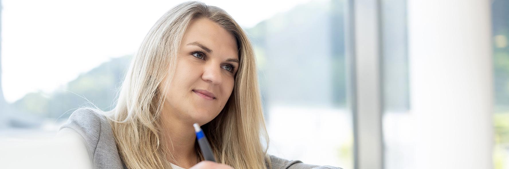 Stephanie sits at a desk with sticky notes and writes on a piece of paper in a bright office environment, while looking in front of her.