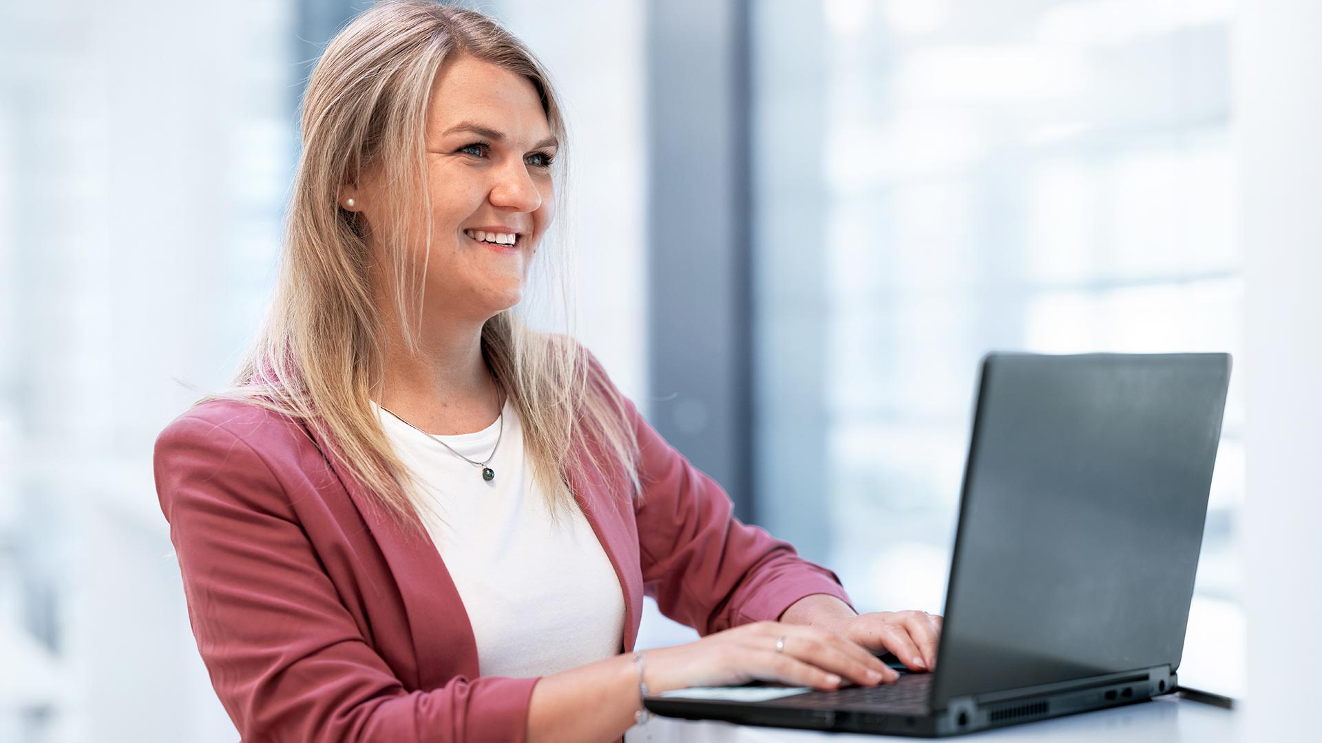 Stephanie sitzt in einem modernen ZEISS Büro und tippt auf ihrem Laptop, während sie lächelnd vor sich hinschaut.