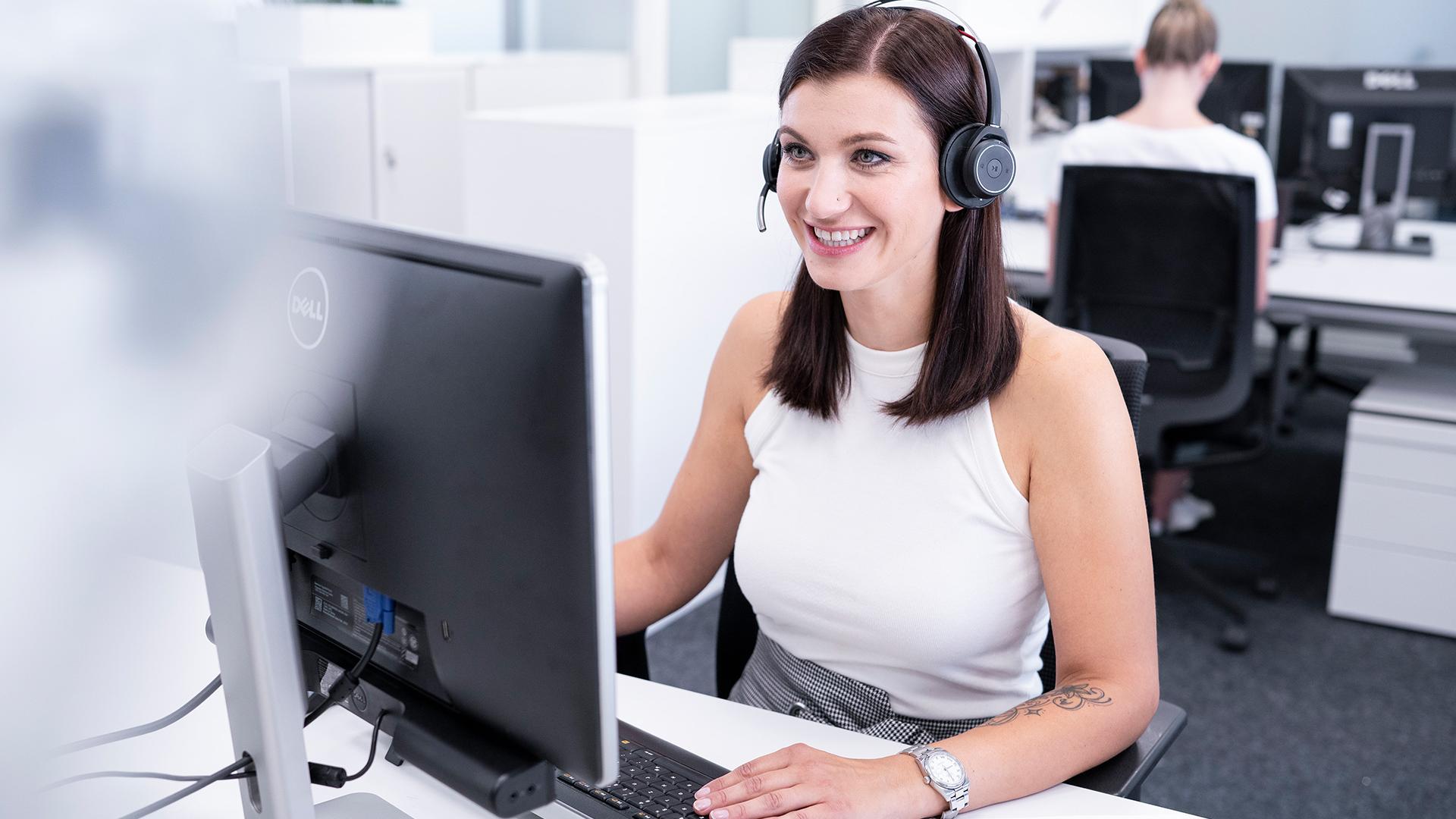 Jenny sitzt im Büro am Schreibtisch und hat ein Headset aufgesetzt