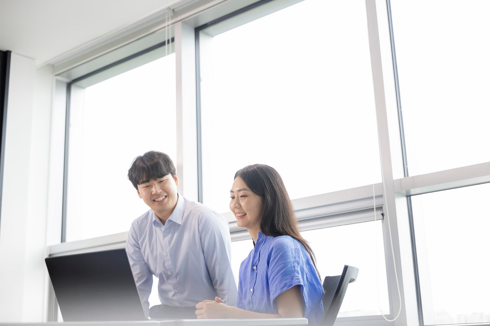 ZEISS Employee JiHye, sitting in office, laughing with a colleague working on laptop