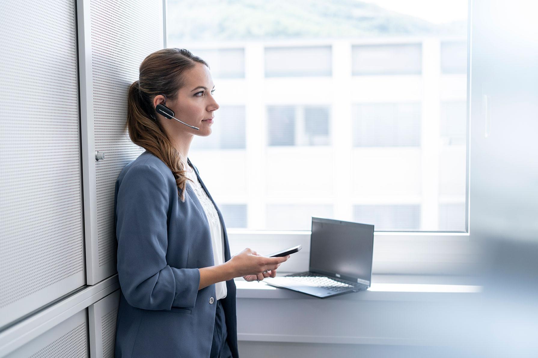 Lisa steht im Büro am Fenster, sie hat Headset an und hält ein Handy in der Hand
