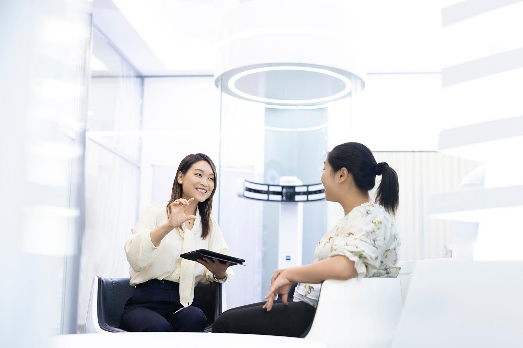 Lizzy chats to a female ZEISS colleague in a bright office while holding a tablet in her left hand.