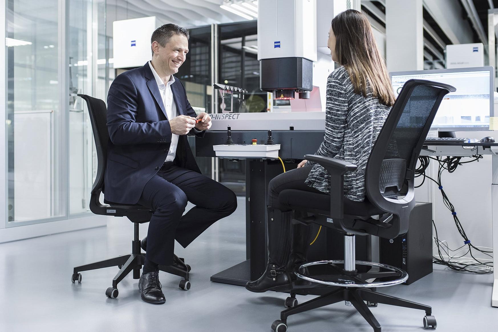 Michael sitzt mit einer Kollegin in einem Raum für Qualitätssicherung. Im Hintergrund steht ein Gerät von ZEISS