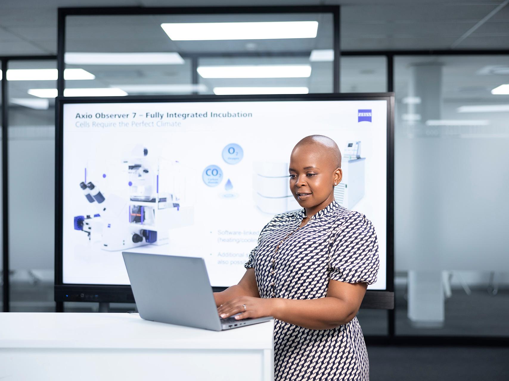 ZEISS employee Thando is working on her laptop in a glass-walled office while a presentation is shown on a digital whiteboard in the background.