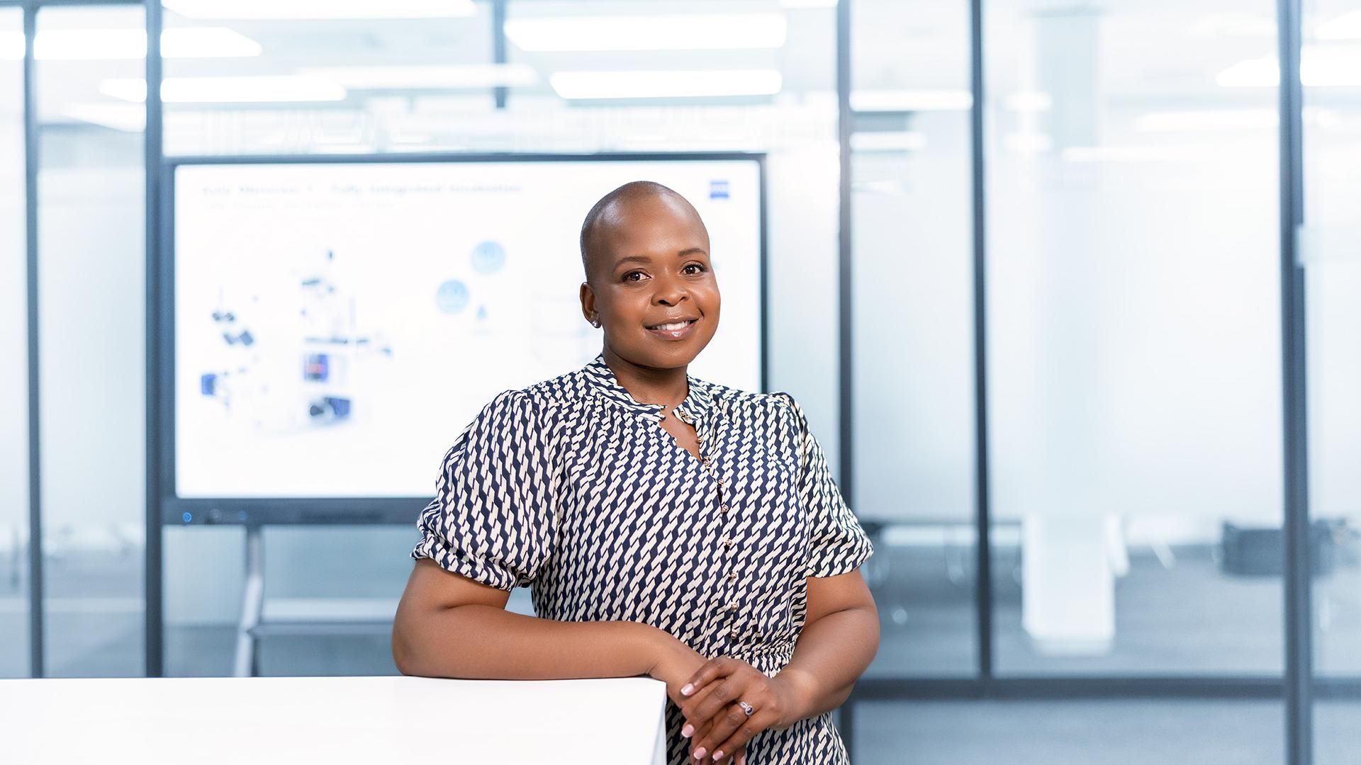 ZEISS employee Thando standing in a glass-walled office, leaning against a table and smiling. A digital projection board with a presentation is shown in the background.