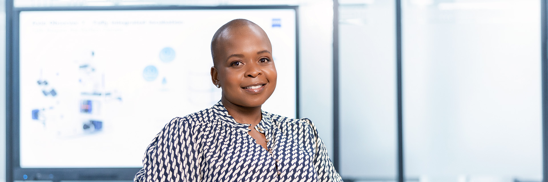 ZEISS employee Thando standing in a glass-walled office, leaning against a table and smiling. A digital projection board with a presentation is shown in the background.