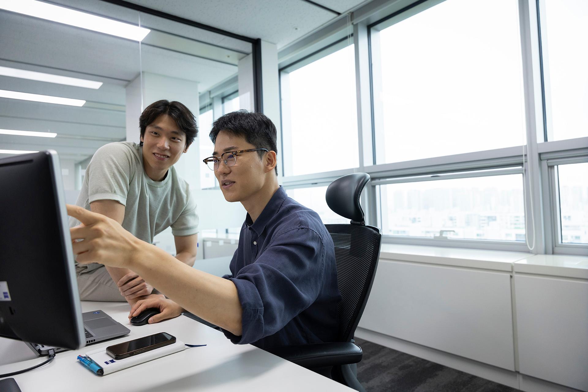 ZEISS employee MinKyue talking to his colleague during both watching a screen in a office near to a big window. 