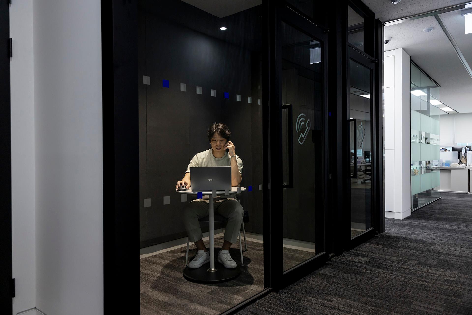 ZEISS employee MinKyue sitting in small single room office, speaking on smartphone and working on a laptop.