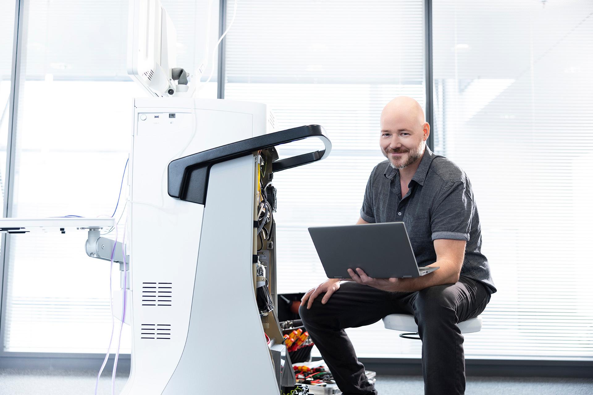 Ralf sits on a swivel stool and holds his laptop in his left hand. He is smiling and facing the camera.