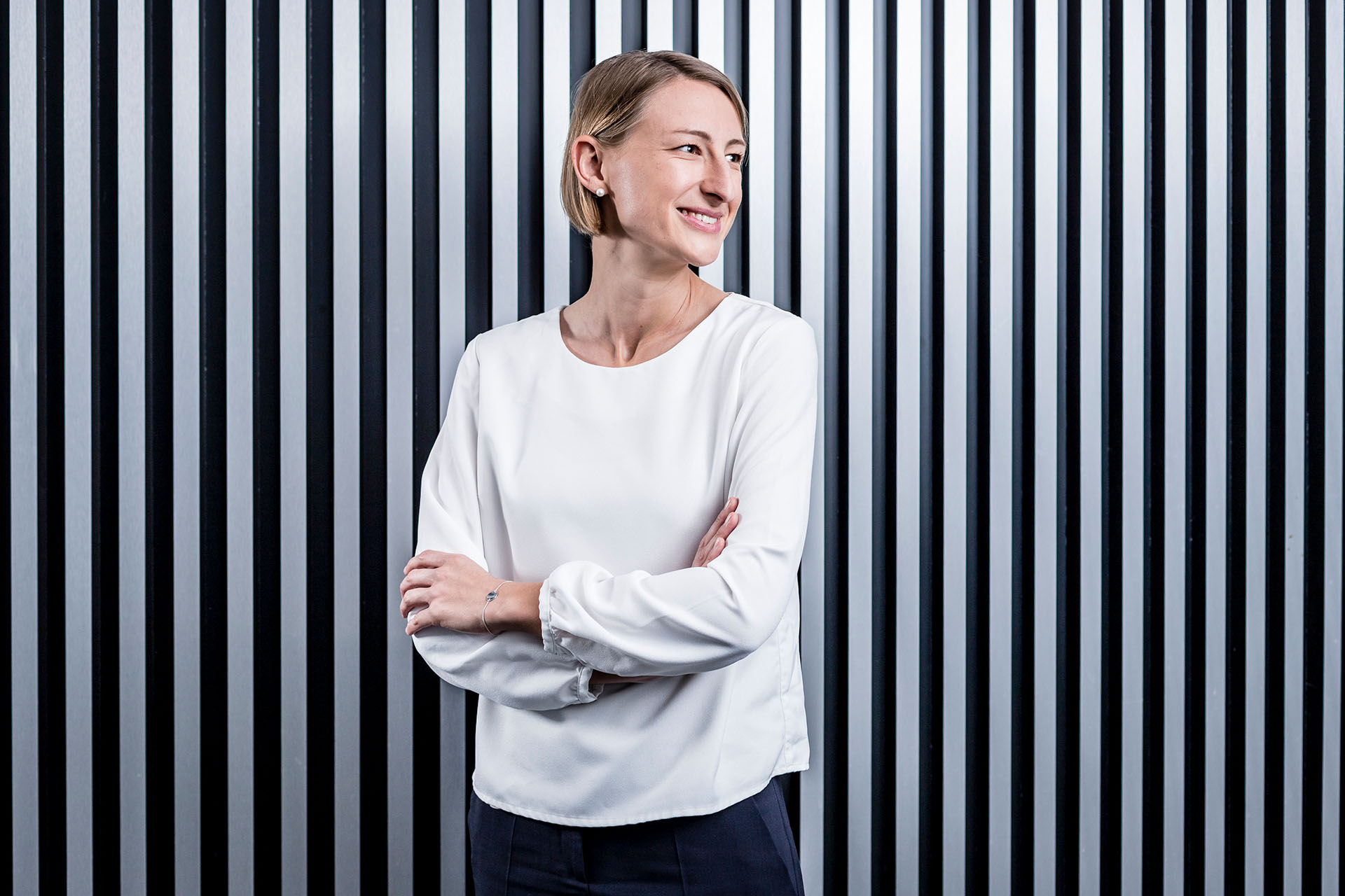 Anna stands in front of a striped panel wall. She looks away from the camera with a friendly expression, while folding her arms.