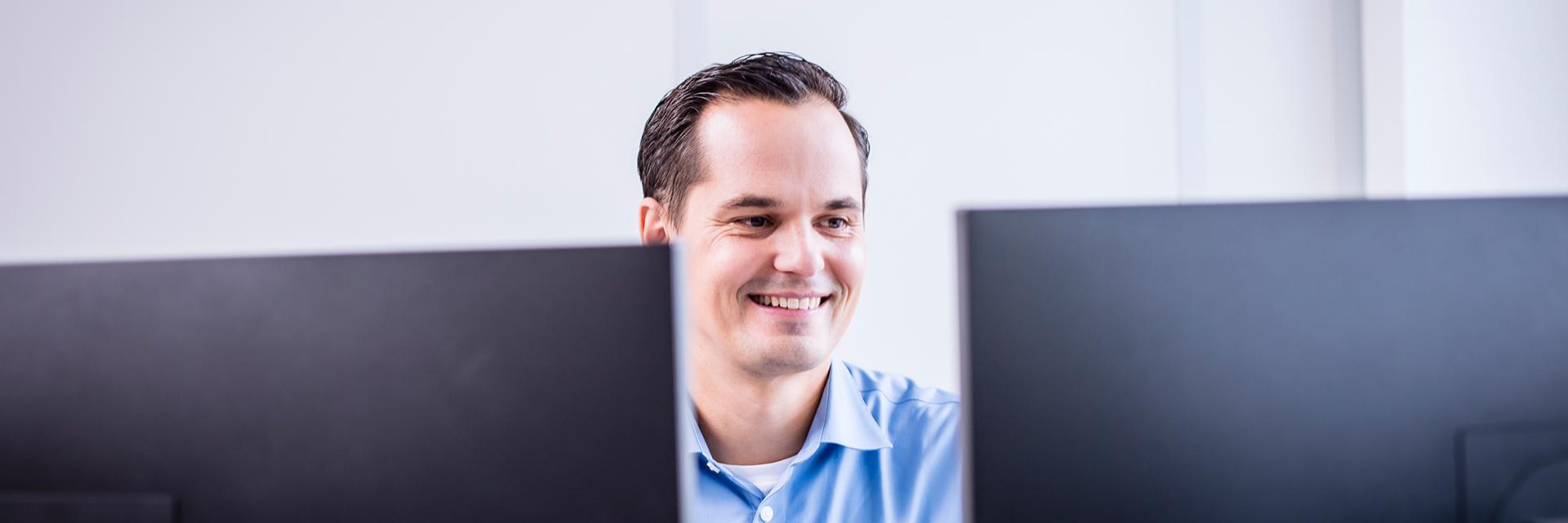 Johannes sitzt vor zwei Bildschirmen, lächelt freundlich und richtet seinen Blick auf einen der Monitore