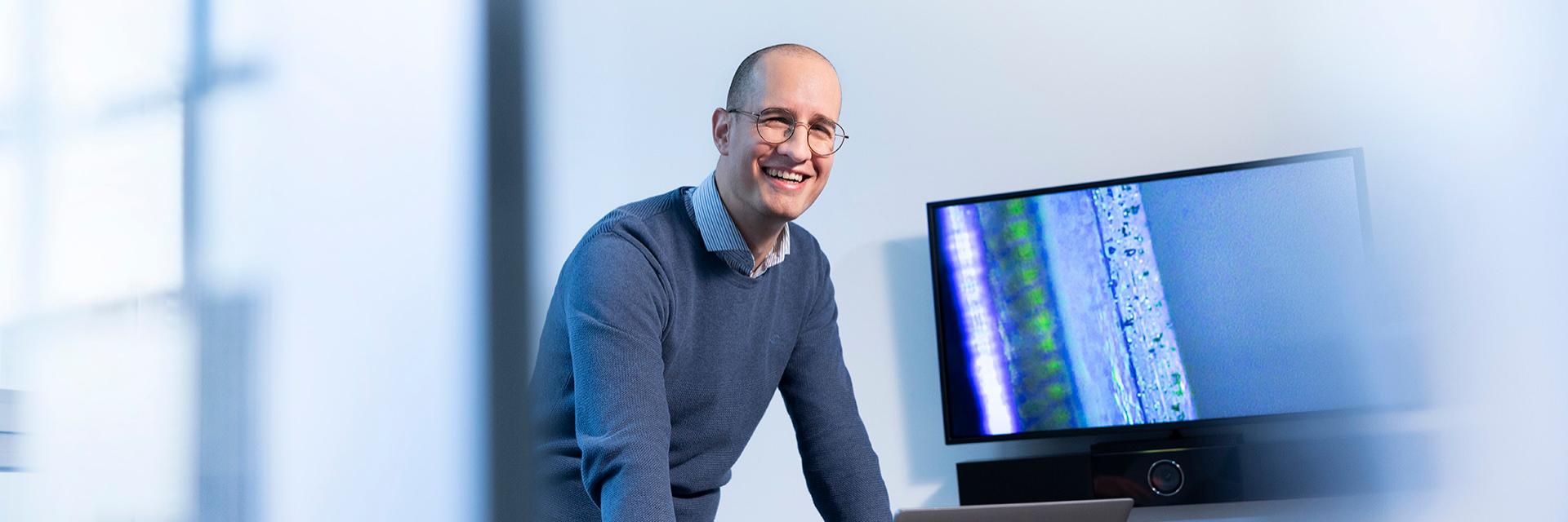 Sam steht lachend am Schreibtisch und arbeitet am Computer in einem separten ZEISS Büro.