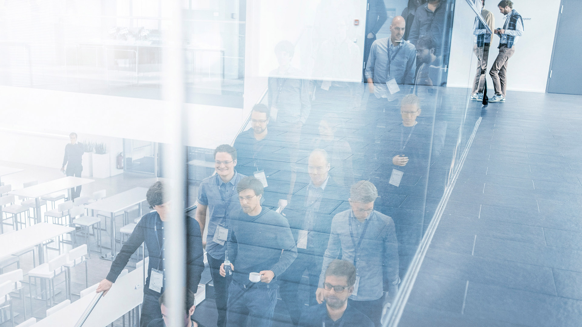 Group of people walks down the stairs. The scene is reflected in a glass partition. A few people wear name badges and hold drinks, which indicates they are in a conference environment. 