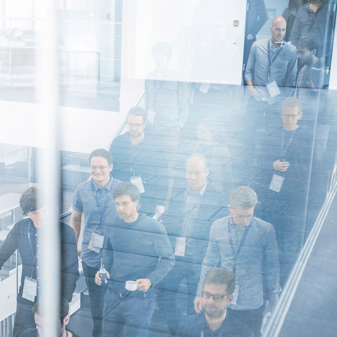 Gruppe von Menschen geht eine Treppe hinunter, die Szene spiegelt sich in einer Glaswand. Einige Personen tragen Namensschilder und halten Getränke, was auf ein Konferenzumfeld hindeutet. 