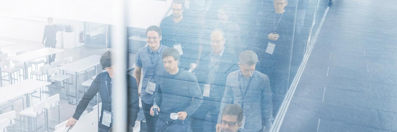 Group of people walks down the stairs. The scene is reflected in a glass partition. A few people wear name badges and hold drinks, which indicates they are in a conference environment. 