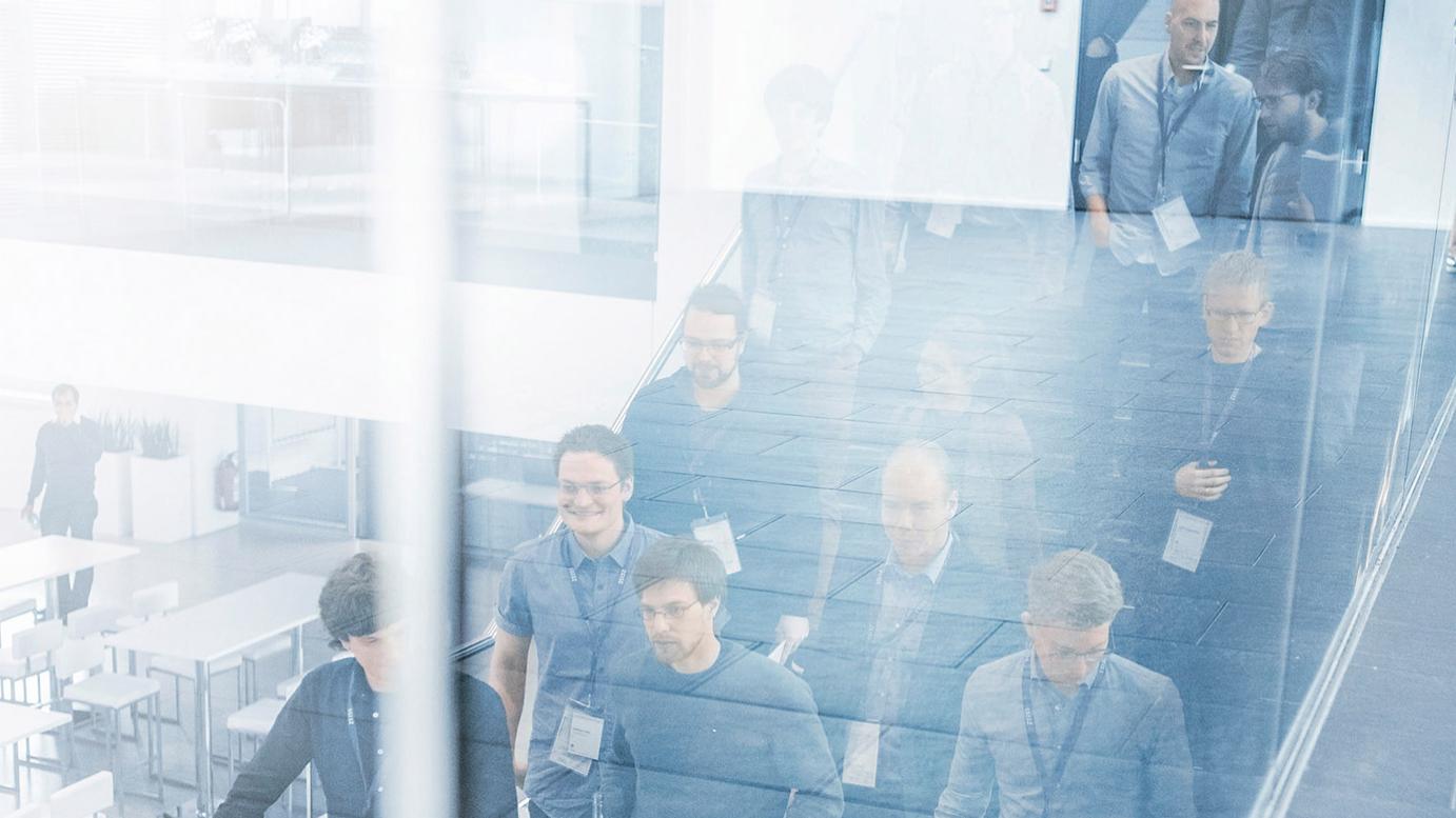 Gruppe von Menschen geht eine Treppe hinunter, die Szene spiegelt sich in einer Glaswand. Einige Personen tragen Namensschilder und halten Getränke, was auf ein Konferenzumfeld hindeutet. 