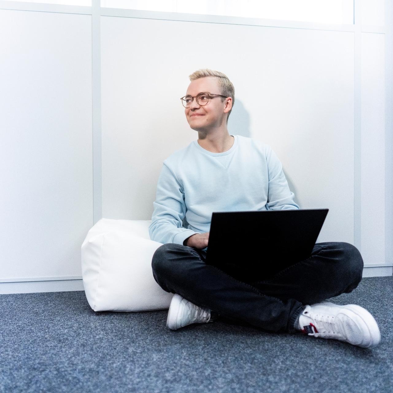 Ein junger Mann sitzt entspannt auf einem Sitzkissen, während er an seinem Laptop arbeitet