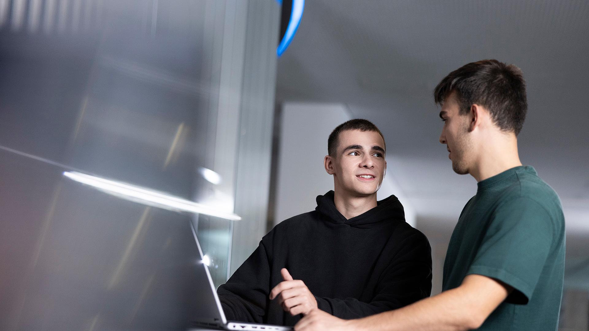 Ein ZEISS Student der Softwaretechnologie blickt seinen Kollegen, gemeinsam an einem Laptop stehend an.