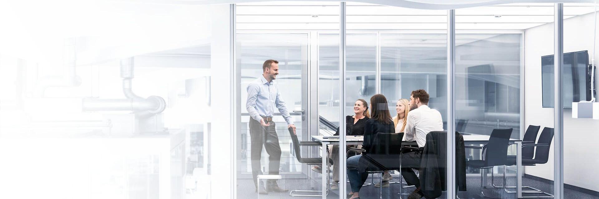 Eine Gruppe von fünf Personen sitzt in einem verglasten Besprechungsraum. Ein Mann steht und gestikuliert, während er spricht.