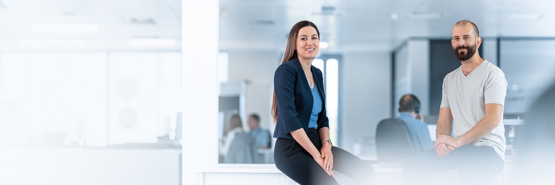 A woman and a man sit in a modern office environment and smile as they look at the camera