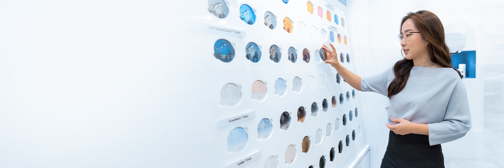 A woman wearing eyeglasses and a light blue blouse touches a display case with colorful glass lenses featuring labels such as 'gradient tints`, 'solid tints' and more.