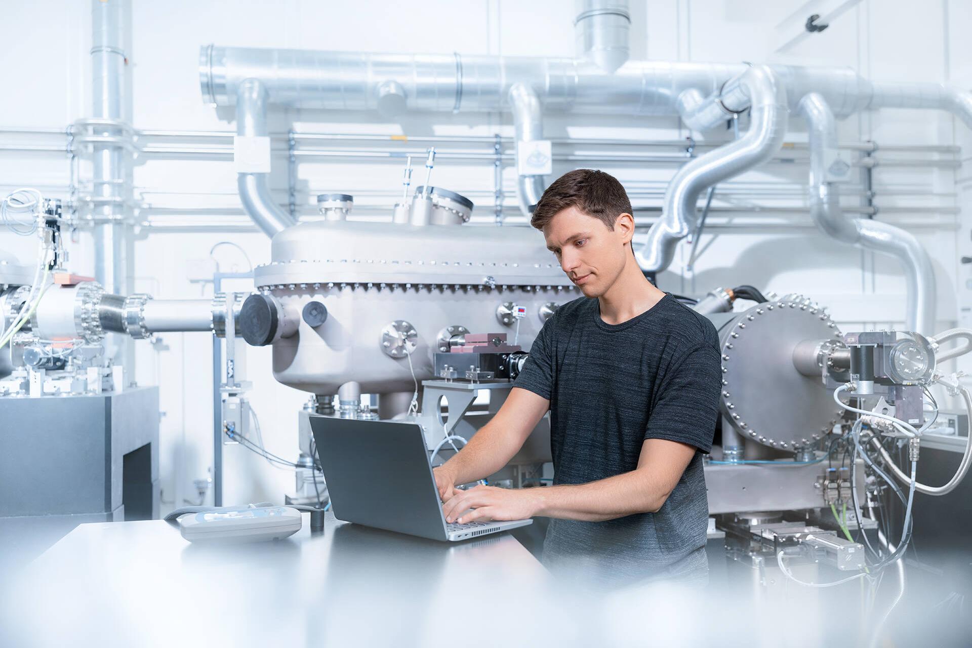 Eine männliche Person steht in einem Produktionsraum und schaut auf einen Laptop