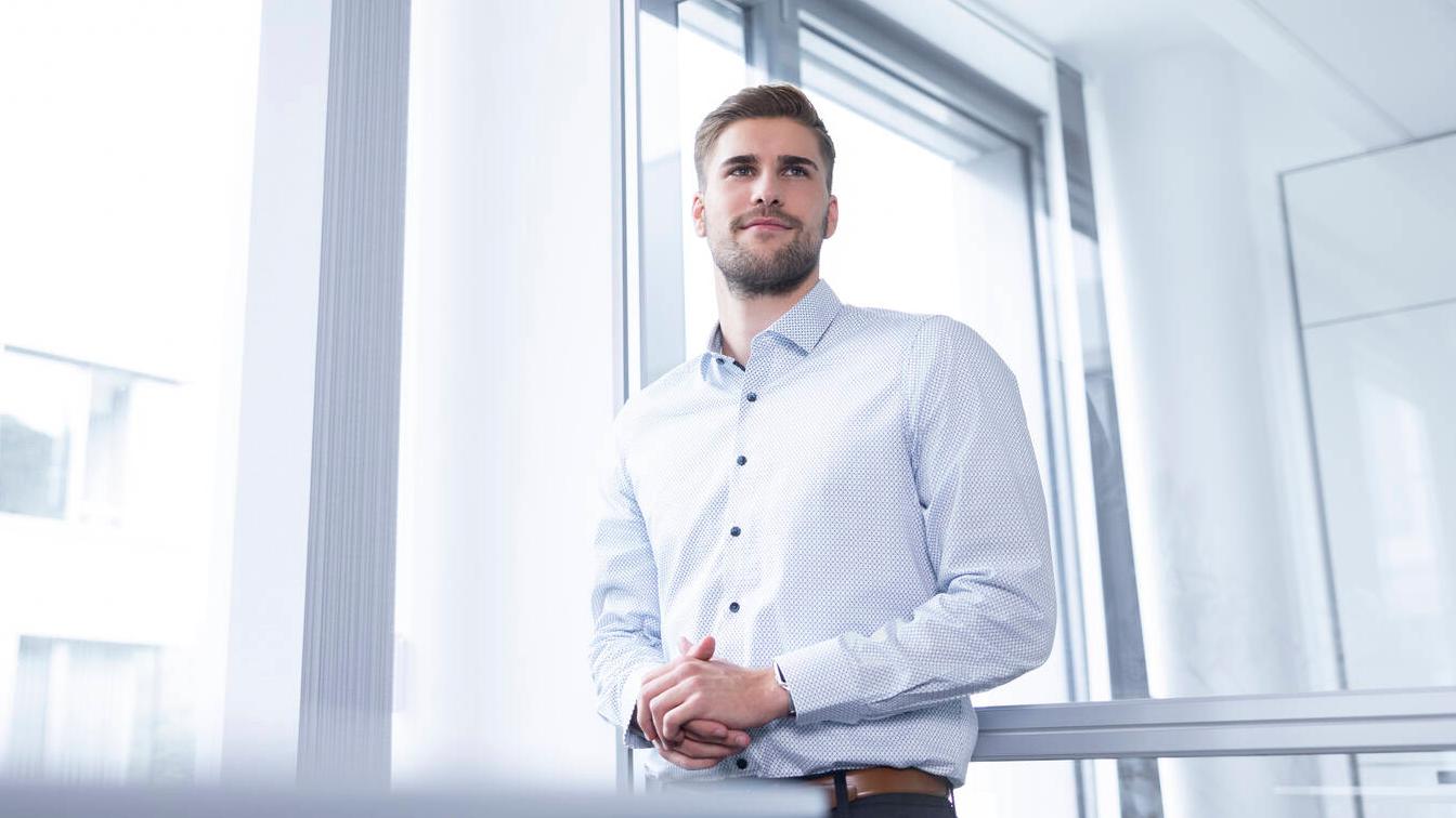 Fabian stehn in einem modernen Büro, er hält die Hände ineinander verschränkt, sein Blick geht an der Kamera vorbei.