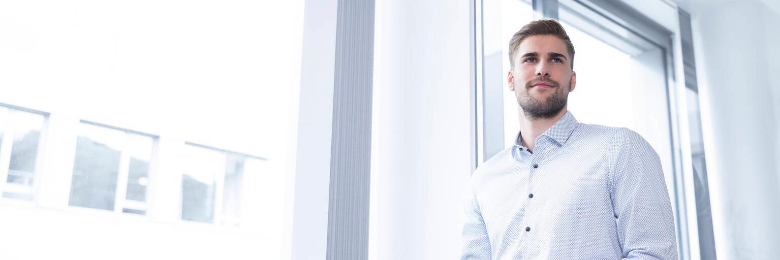 Fabian stehn in einem modernen Büro, er hält die Hände ineinander verschränkt, sein Blick geht an der Kamera vorbei.