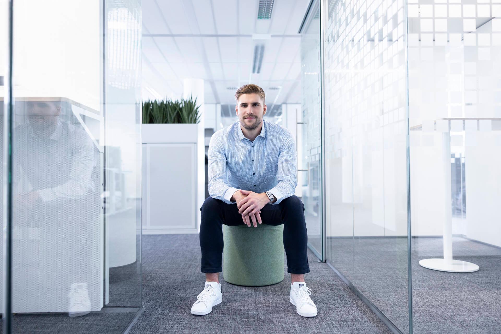 Fabian sitzt nach vorne gebeugt auf einem Hocker im Büro mit Glaswänden.